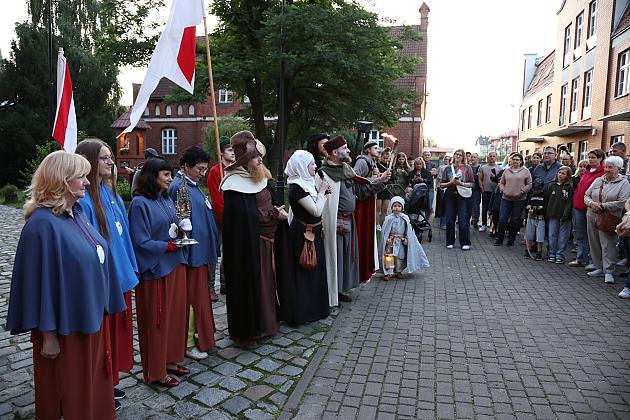 Orszak ludzi w historycznych strojach z flagami na ulicy podczas jarmarku.