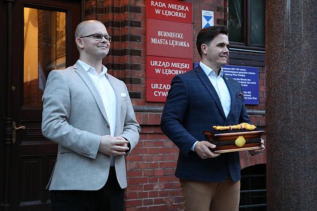 Dwaj mężczyźni stoją przy ceglanej ścianie, jeden trzyma relikwiarz świętego Jakuba.