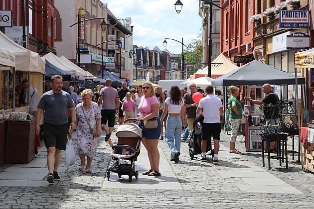 Słoneczny dzień na jarmarku z tłumem ludzi, straganami i kamienicami.